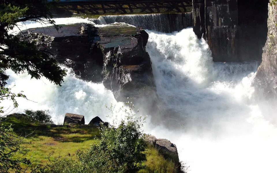 A powerful waterfall cascades over dark, jagged rocks, with mist rising and green grass in the foreground.