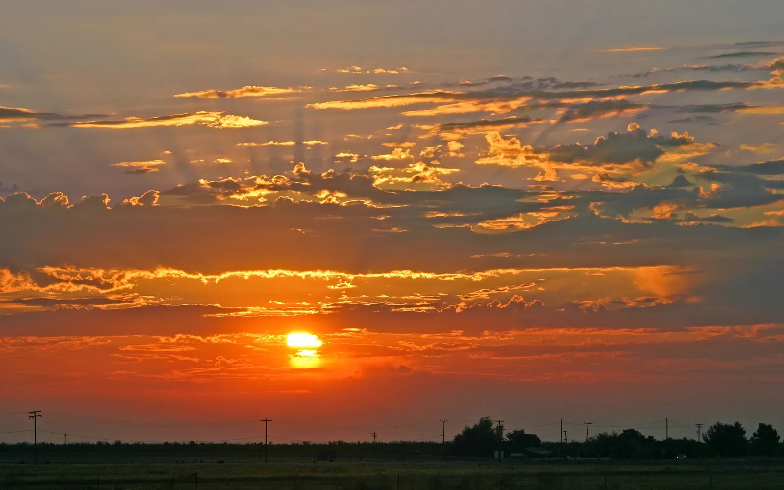 The sun sets over a flat landscape, casting golden rays through dramatic clouds.
