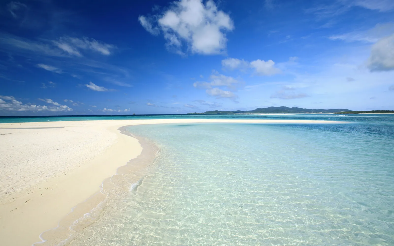 A serene white sand beach curves into exceptionally clear ocean water transitioning to deeper blue hues.