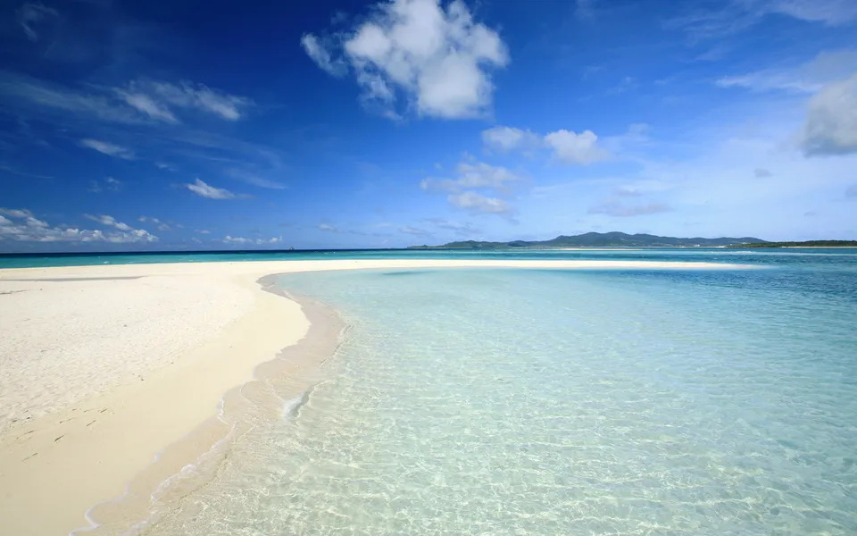 A serene white sand beach curves into exceptionally clear ocean water transitioning to deeper blue hues.