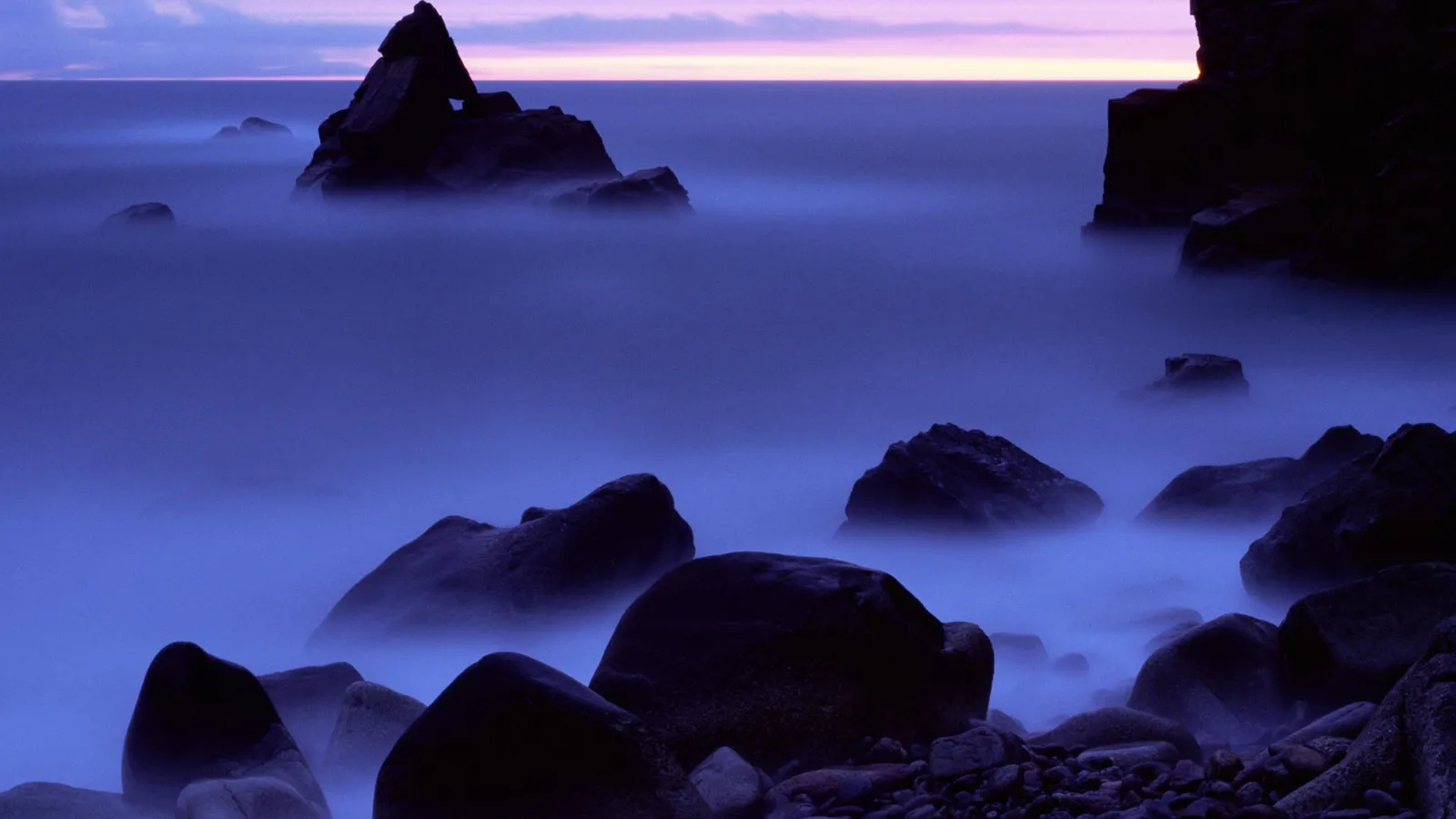 Jagged dark rocks emerge from a thick purple mist at dawn on a rocky coastline.