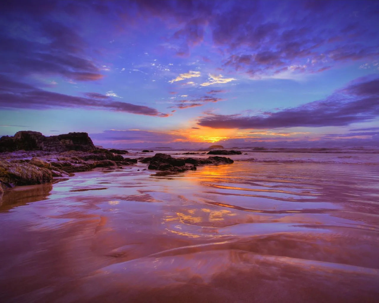 A vibrant sunset with purple and orange hues paints the sky over a rocky shoreline and wet sand reflecting the colors.