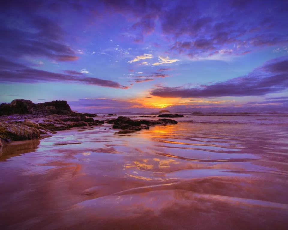 A vibrant sunset with purple and orange hues paints the sky over a rocky shoreline and wet sand reflecting the colors.