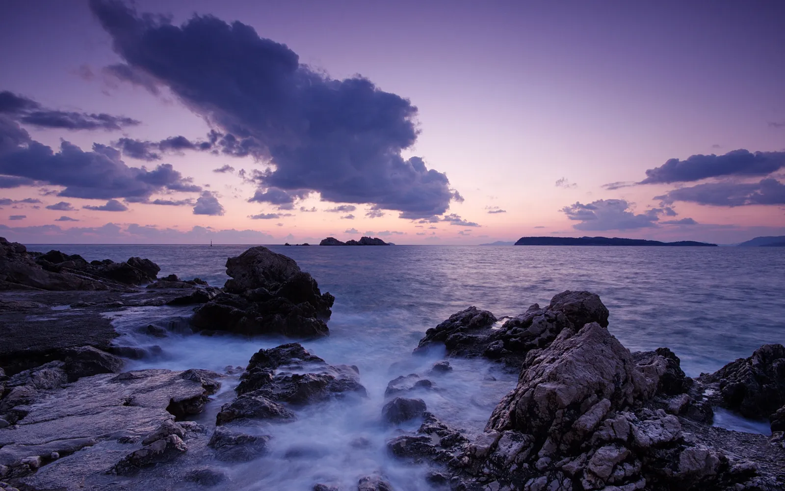 A serene sunset with purple and pink hues over a rocky coastline and crashing waves.