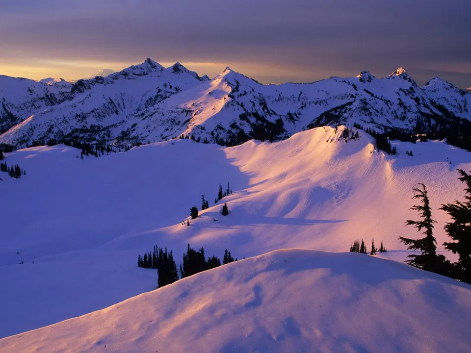 Snow-covered mountain ridges illuminated by the soft, warm hues of the setting sun with purple sky.