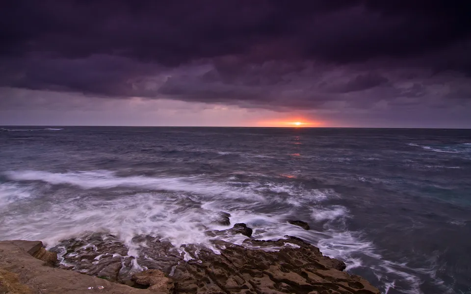 A dramatic purple sunset over a dark, rocky coastline with waves crashing.