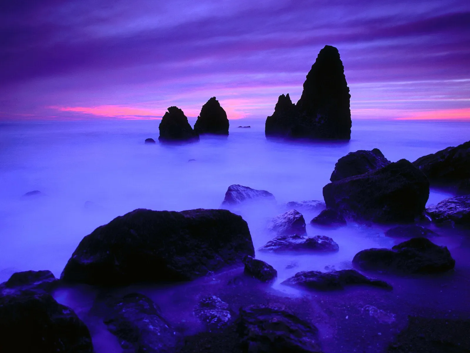 Dark, jagged rocks emerge from misty, purple-hued ocean water during a vibrant sunset.