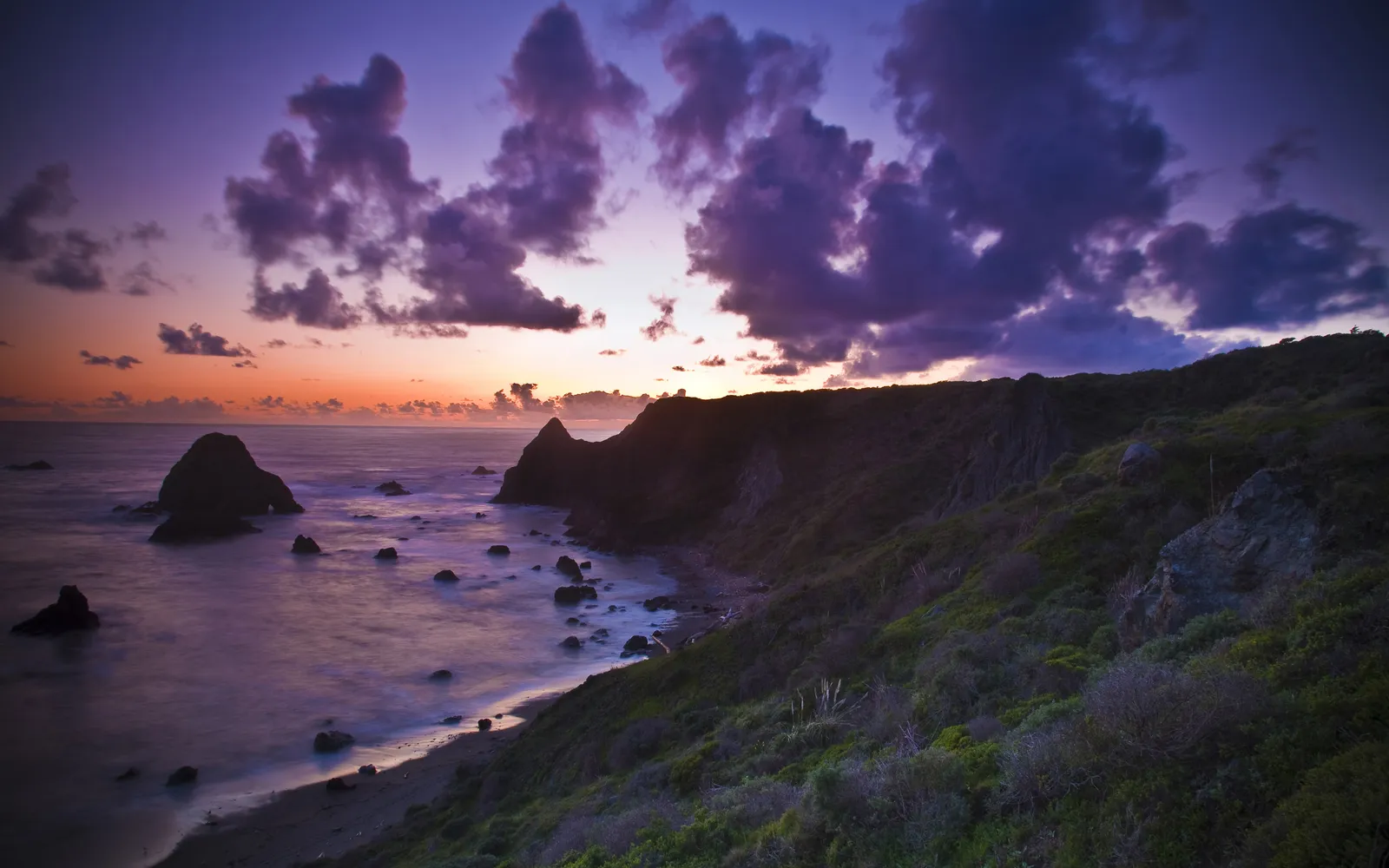 A vibrant sunset with purple clouds over rugged coastal cliffs and the ocean.