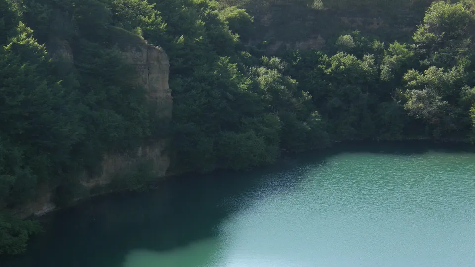 A tranquil turquoise quarry lake is bordered by a steep, tree-covered cliff face.