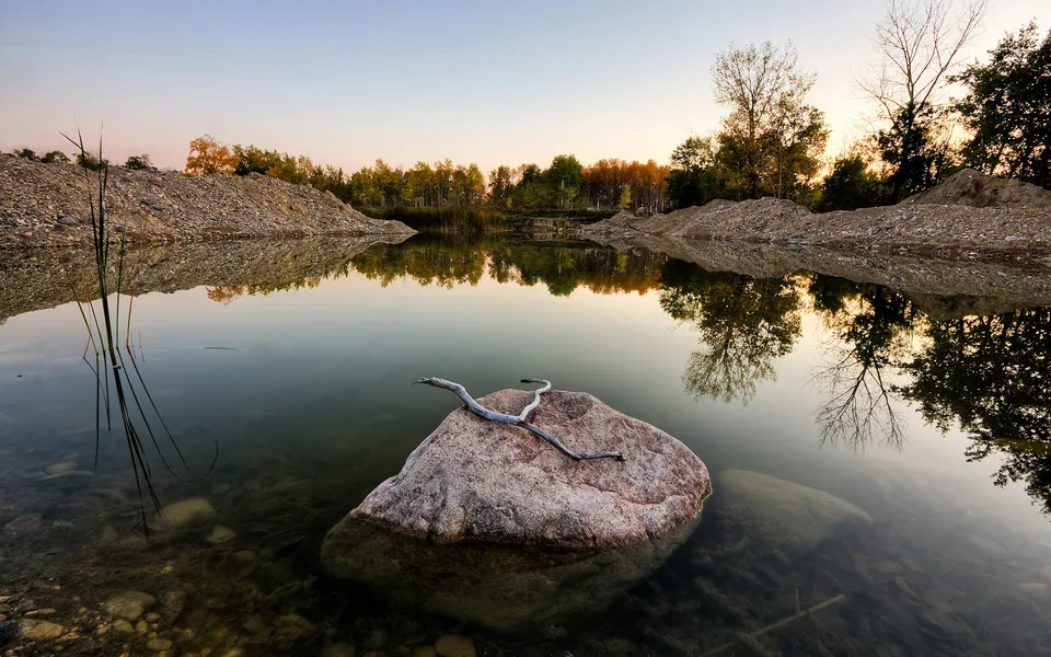 A large rock with a branch rests in a calm quarry lake reflecting trees and a soft sky at sunset.