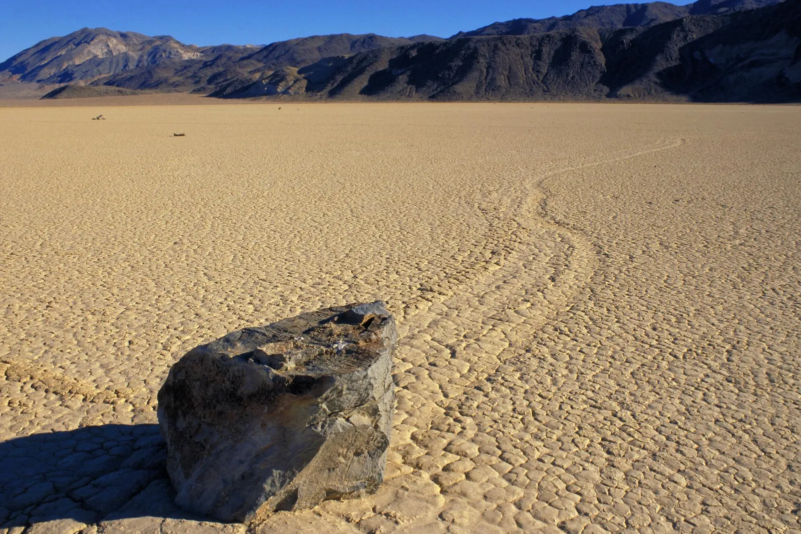 A large rock sits in the foreground on a cracked, dry desert playa with mountains in the background and a faint trail leading away.