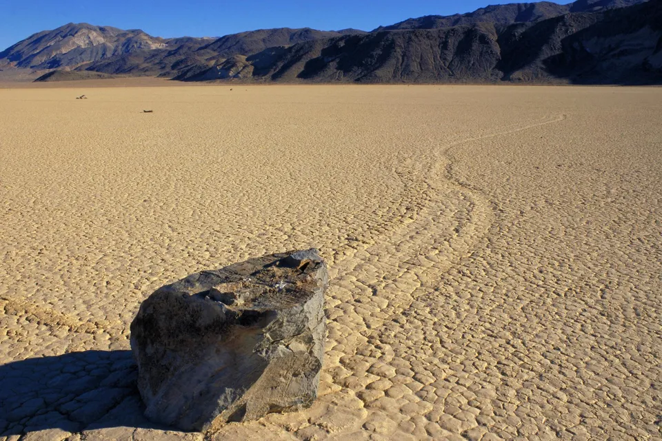 A large rock sits in the foreground on a cracked, dry desert playa with mountains in the background and a faint trail leading away.