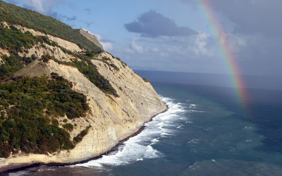 A vibrant rainbow arcs across a cloudy sky above a steep, rocky coastline with waves crashing on the shore.