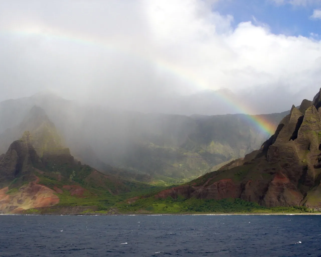 A vibrant rainbow arcs across a misty sky over rugged, green-covered coastal mountains.