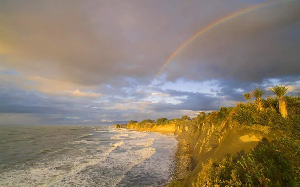 A rainbow arcs across a cloudy sky over a rugged coastline with waves crashing on the shore.