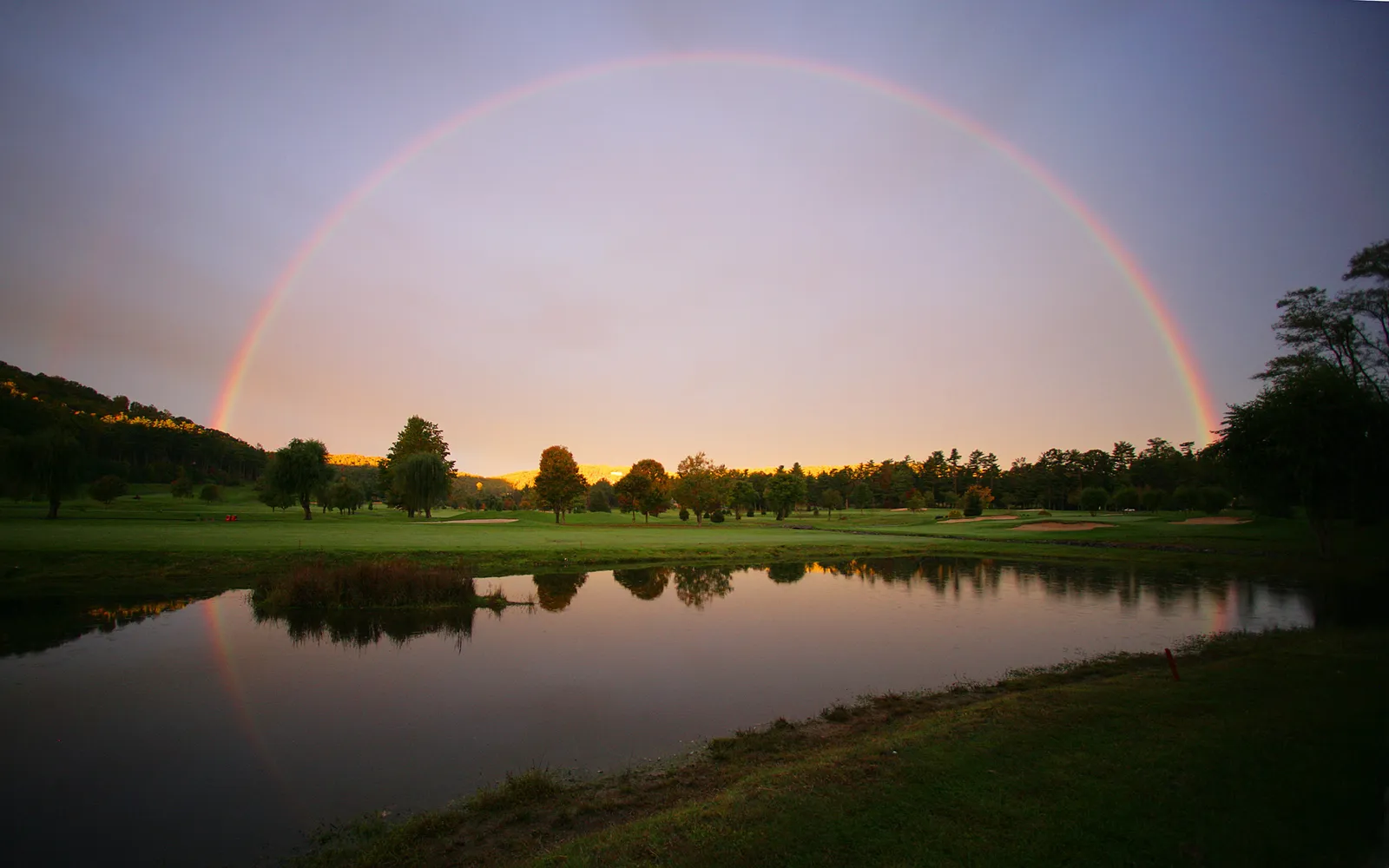 A full rainbow arcs across a pastel sky over a golf course with a pond in the foreground.