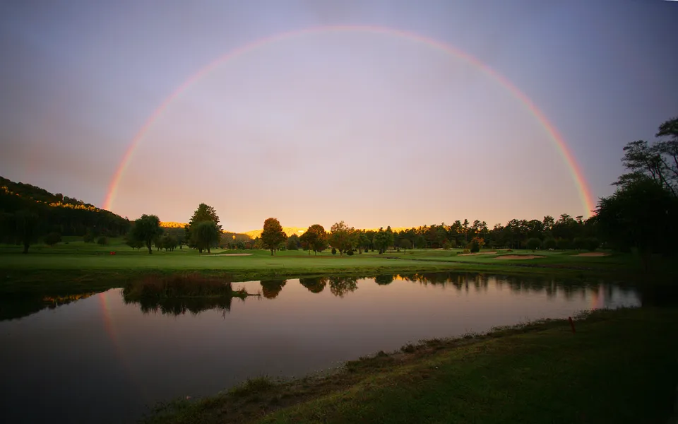A full rainbow arcs across a pastel sky over a golf course with a pond in the foreground.