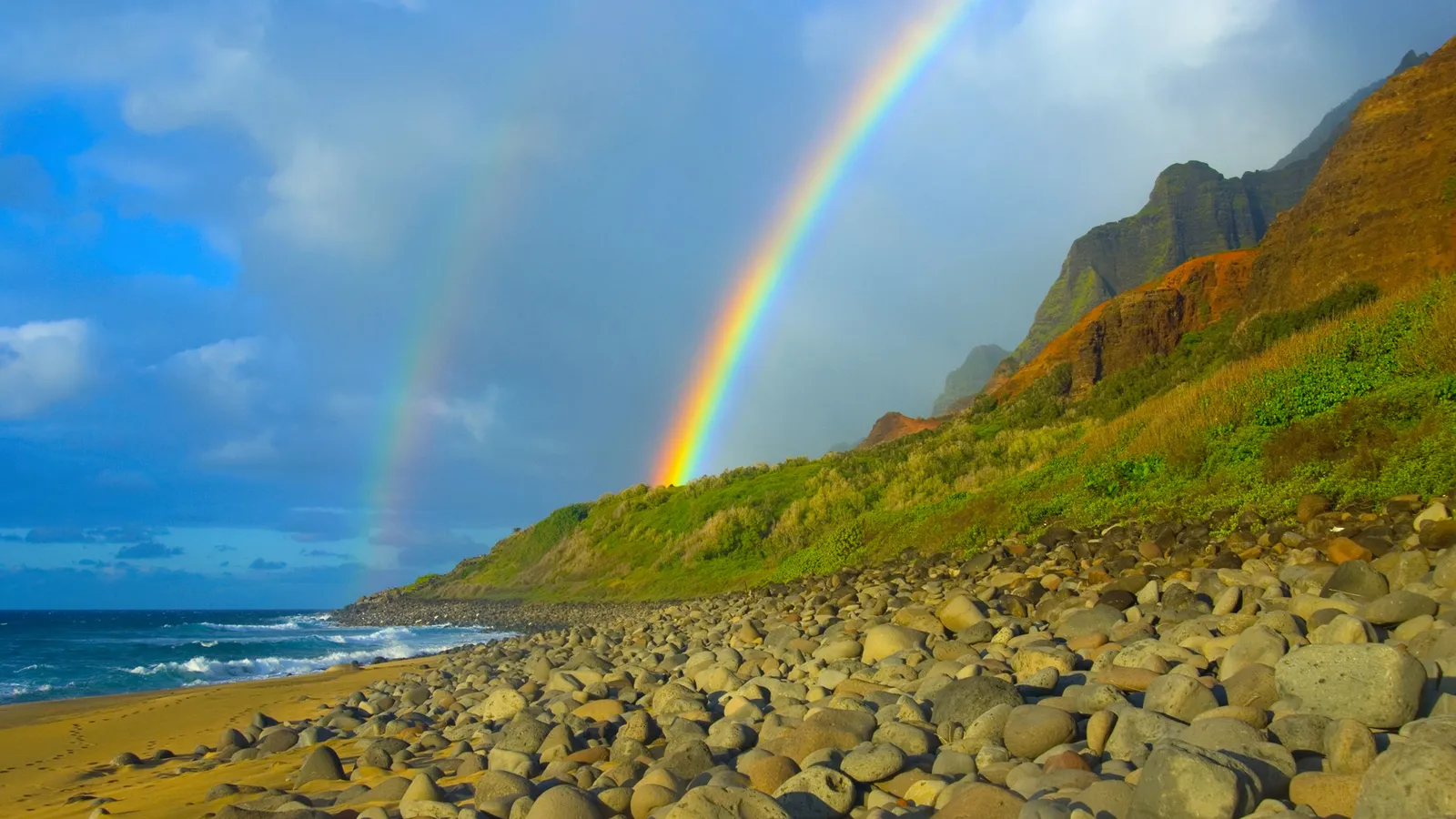 A double rainbow arches over a lush green cliff face next to a rocky beach and the ocean.