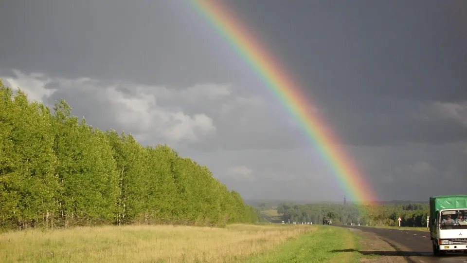 A bright rainbow arcs across a stormy sky over a grassy field and a road with a truck.