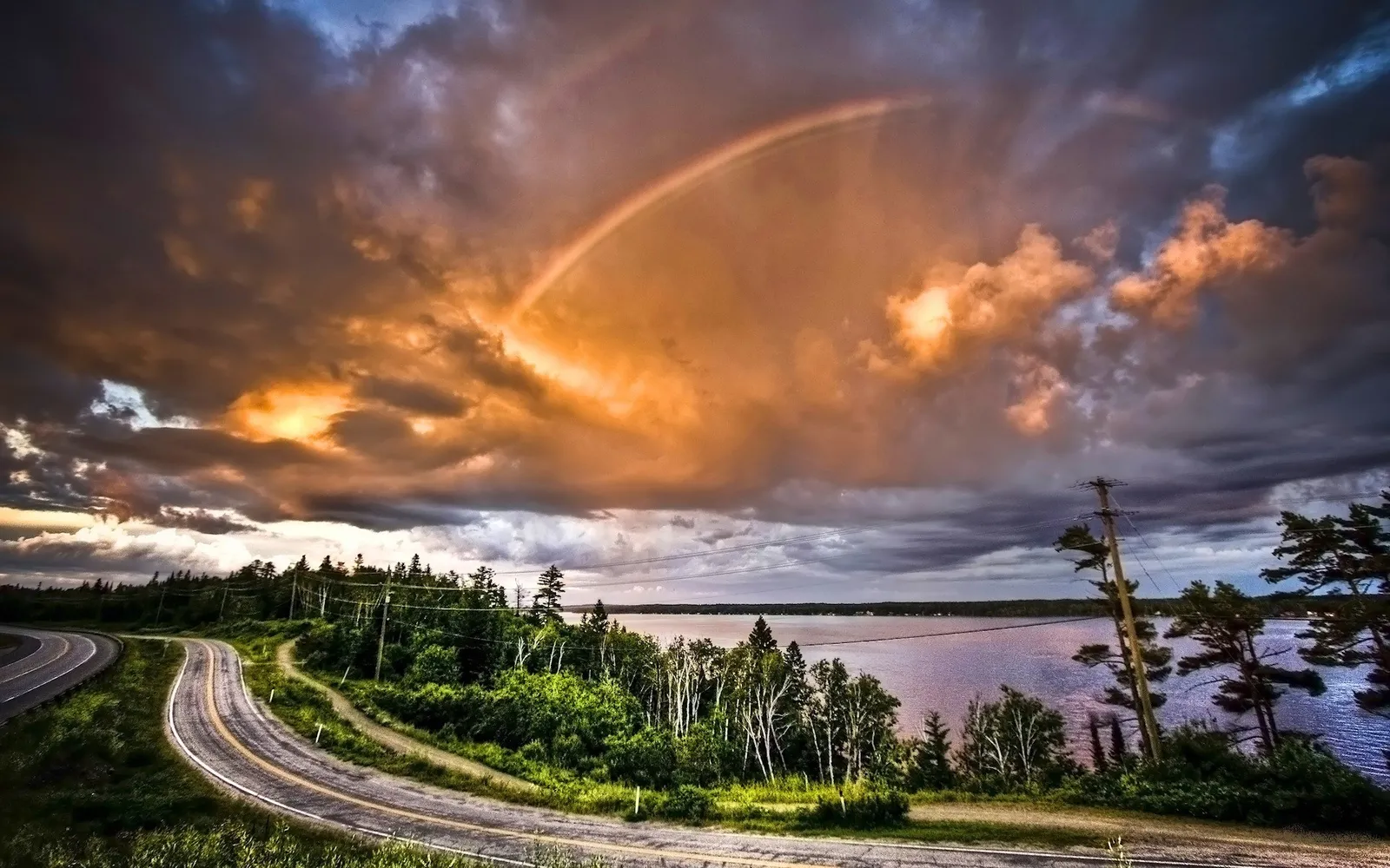 A vibrant rainbow arcs across a dramatic sky filled with orange and dark clouds, above a calm lake and tree-lined shore.