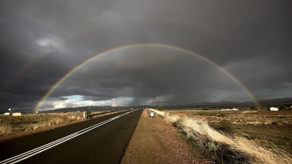 A double rainbow spans across a dark, stormy sky over a long, empty road.