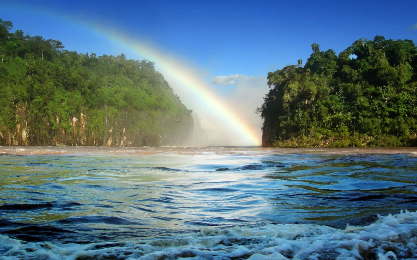 A vibrant rainbow arcs over a misty waterfall between two lush green cliffs, with turbulent water in the foreground.