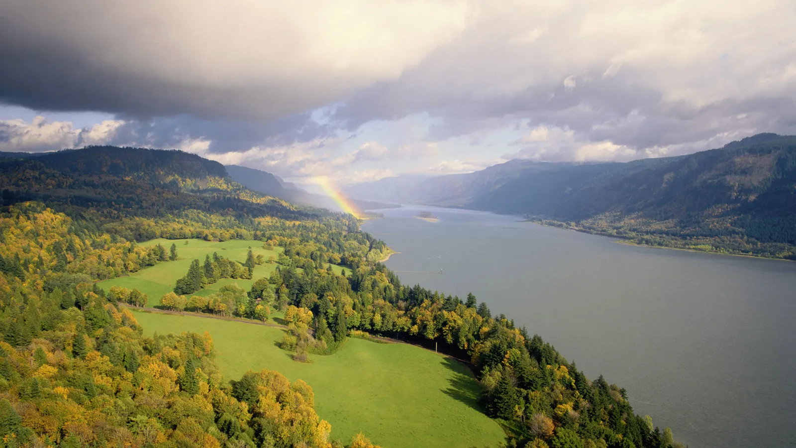 An aerial view of a wide river flowing through a valley with green fields and autumn trees, a rainbow arcs in the sky.