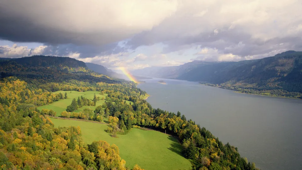 An aerial view of a wide river flowing through a valley with green fields and autumn trees, a rainbow arcs in the sky.