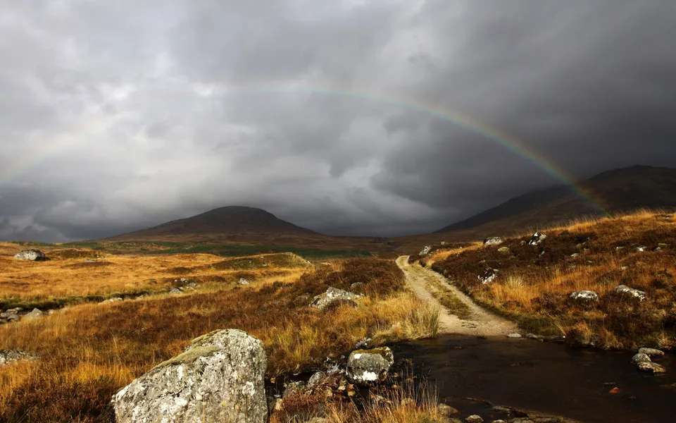 A dirt track winds through a golden, grassy highland landscape under a dramatic sky with a rainbow.
