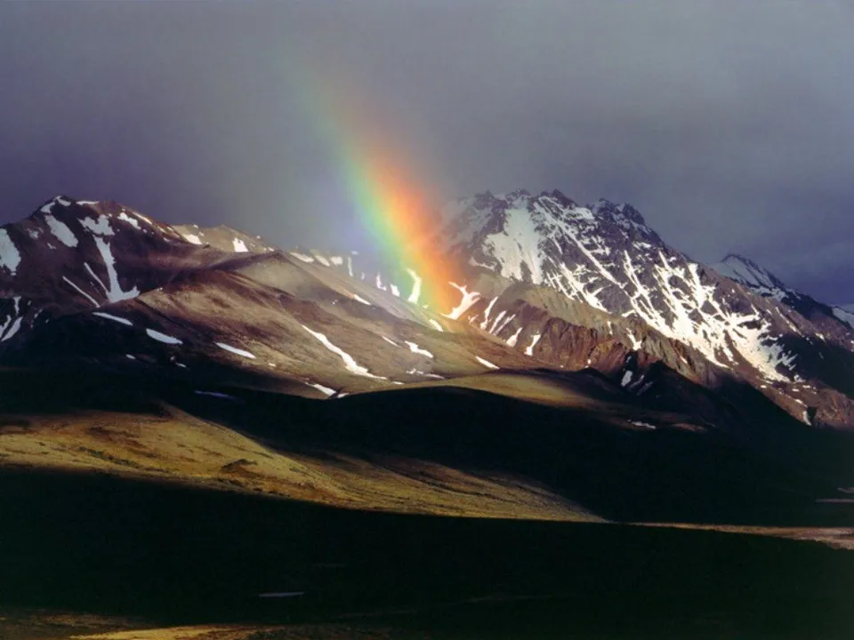 A vibrant rainbow arcs across a dark sky over rugged, snow-dusted mountain peaks.