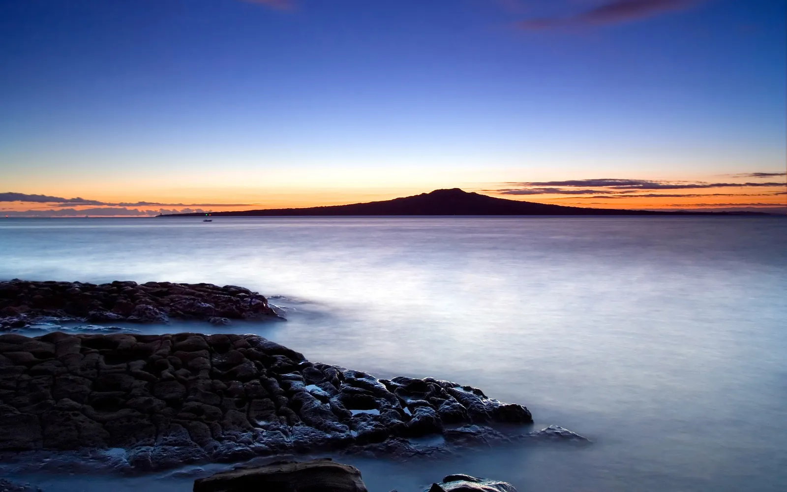 A silhouette of Rangitoto Island is visible across a calm sea at sunset, with rocky foreground.