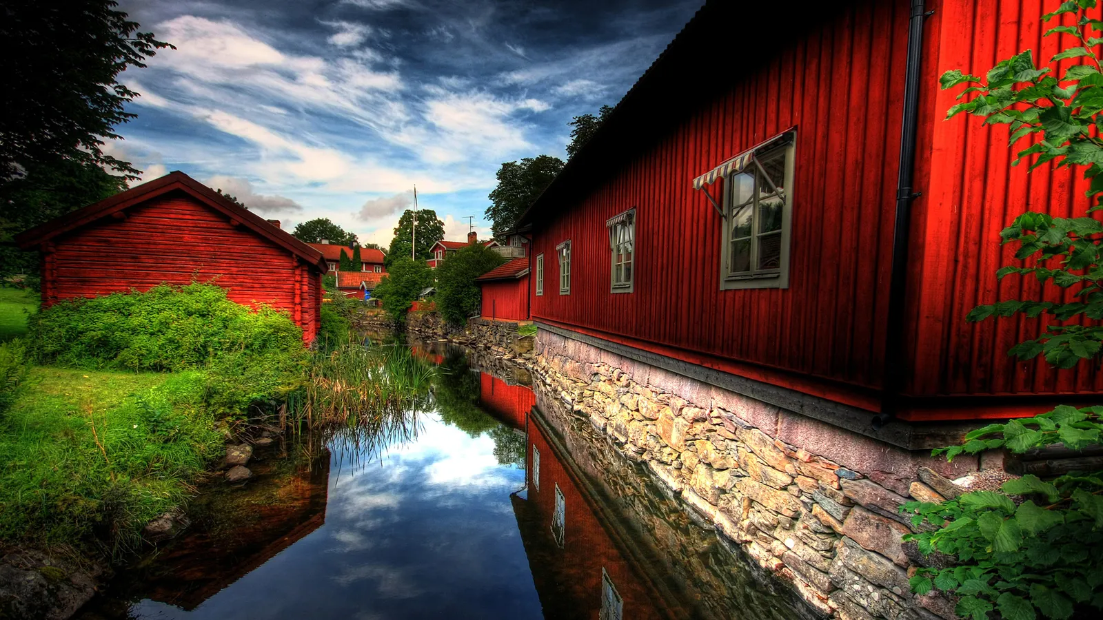 Two traditional red wooden buildings with stone foundations reflected in a calm body of water under a cloudy sky.