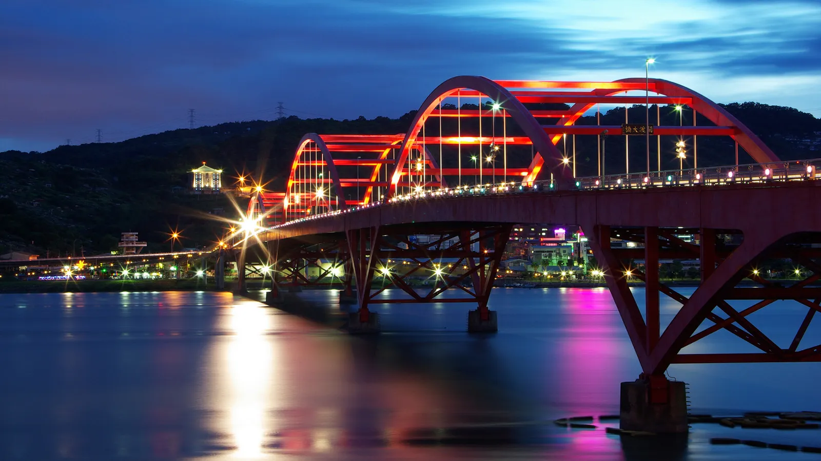 A red arched bridge illuminated with lights over a body of water at dusk, with reflections on the water.