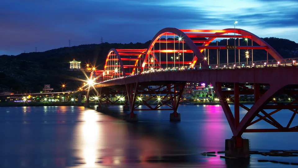 A red arched bridge illuminated with lights over a body of water at dusk, with reflections on the water.