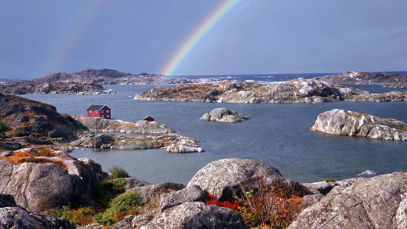 A small red house sits on a rocky island in the sea, with a rainbow arching across the sky.