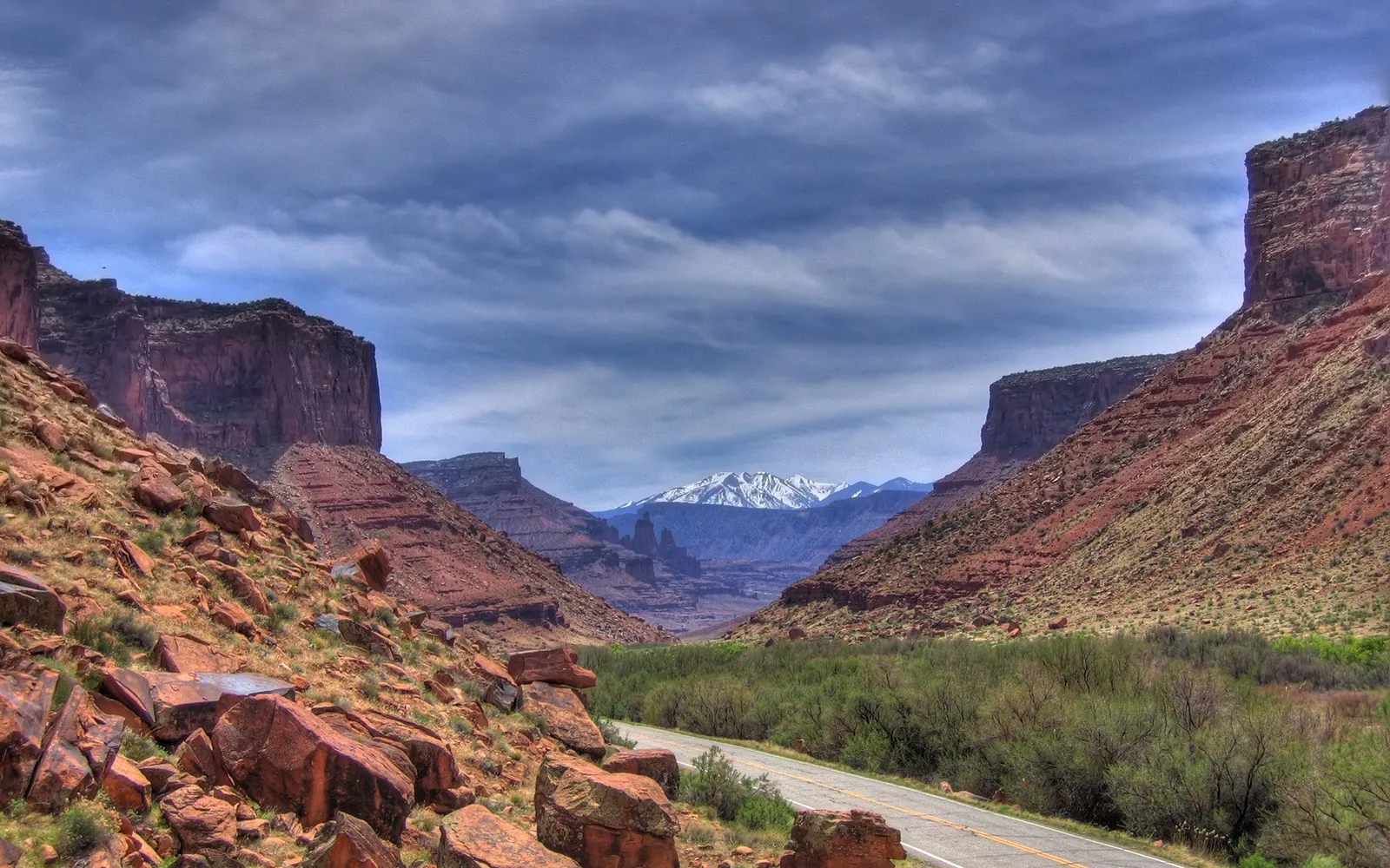 A paved road winds through a red rock canyon towards distant snow-capped mountains under a cloudy blue sky.