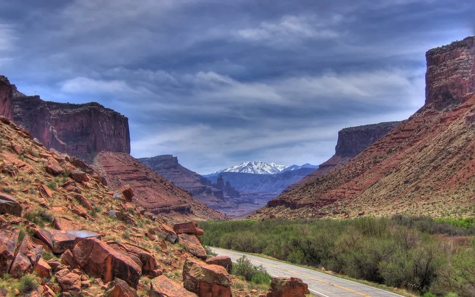 A paved road winds through a red rock canyon towards distant snow-capped mountains under a cloudy blue sky.