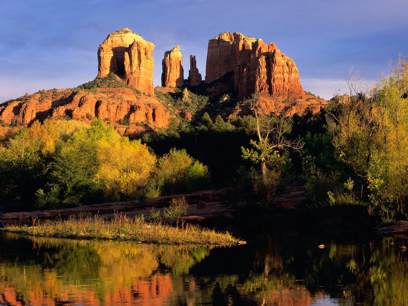 Iconic red rock formations stand tall, with trees in autumn colors and their reflections in a calm body of water.