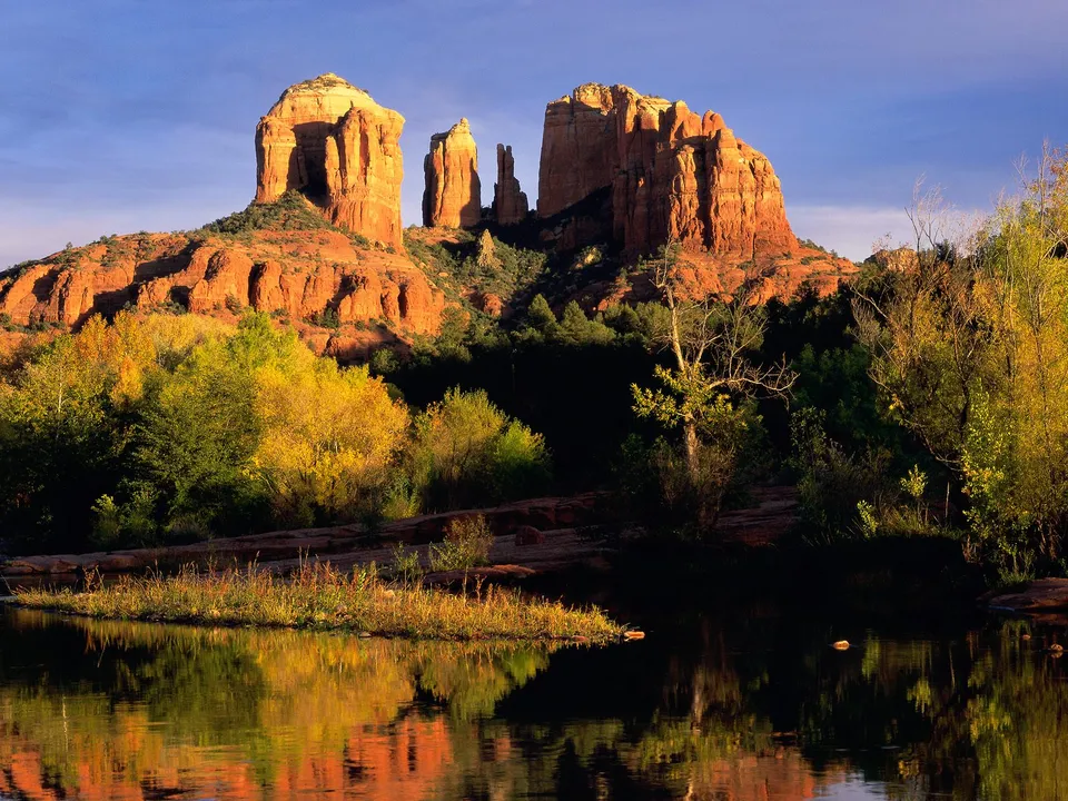 Iconic red rock formations stand tall, with trees in autumn colors and their reflections in a calm body of water.