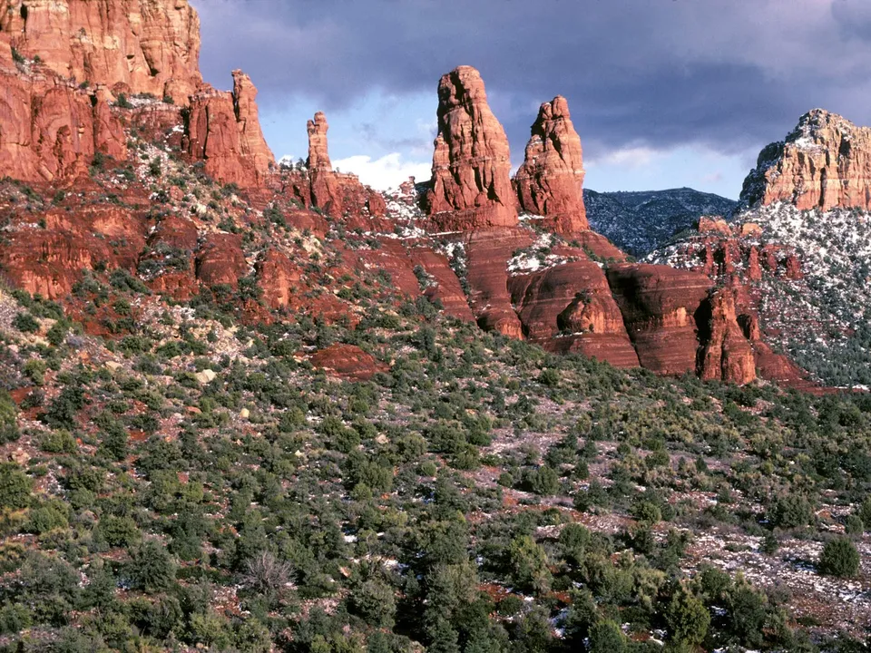 Towering red rock formations with patches of snow, under a dark, cloudy sky.