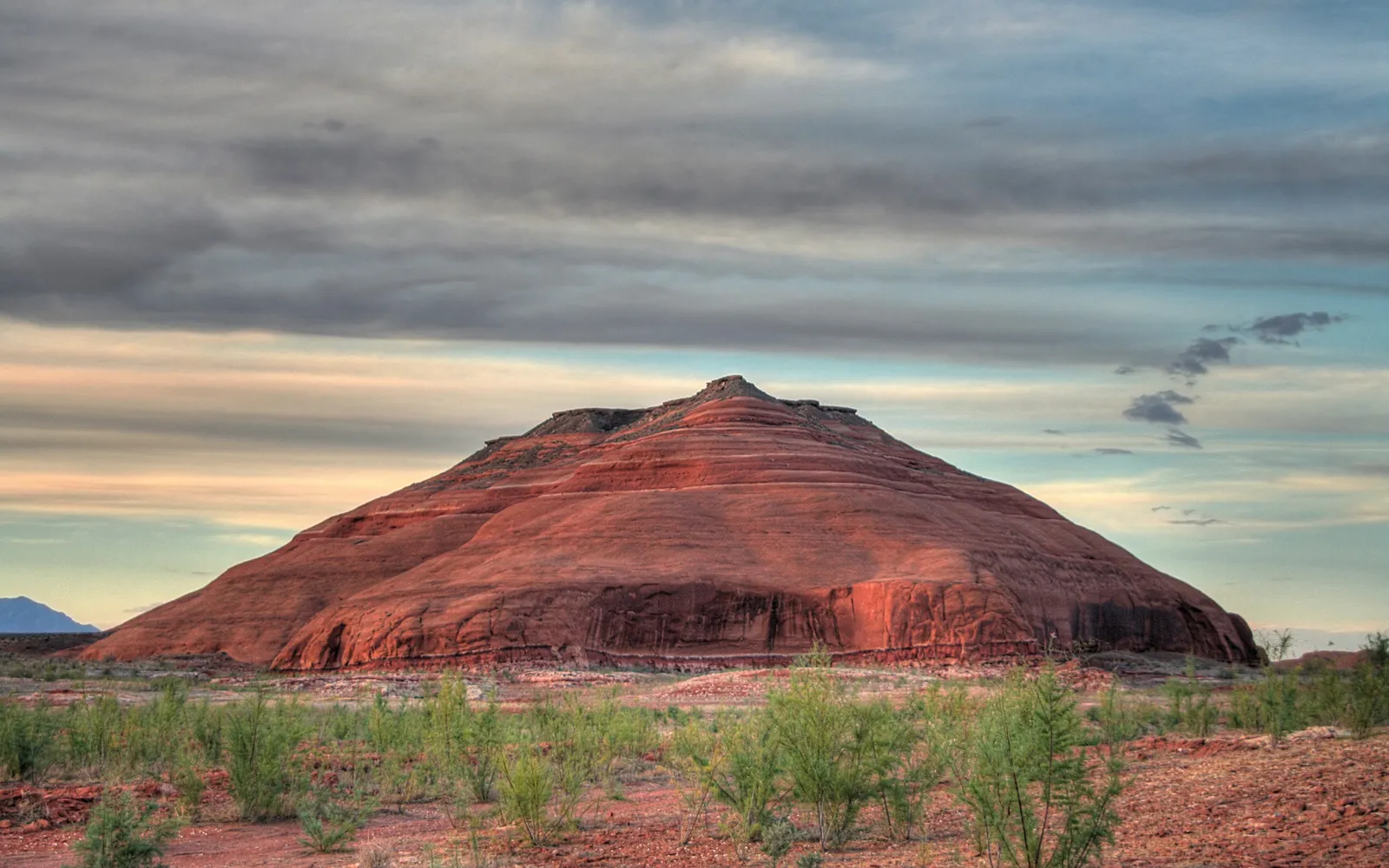 A large, layered red rock mountain dominates the landscape under a cloudy sky with streaks of color.