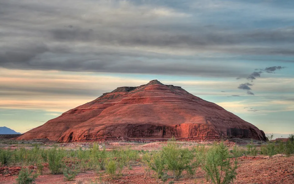 A large, layered red rock mountain dominates the landscape under a cloudy sky with streaks of color.