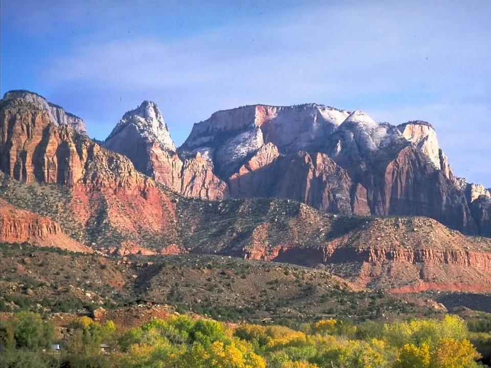 A wide view of towering red rock mountains with patches of snow on top, under a clear blue sky.