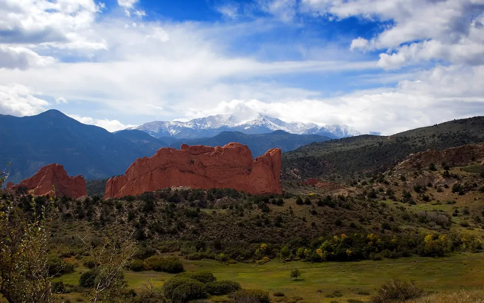 A vast landscape featuring prominent red rock formations in the foreground with snow-capped mountains in the background under a blue sky with clouds.
