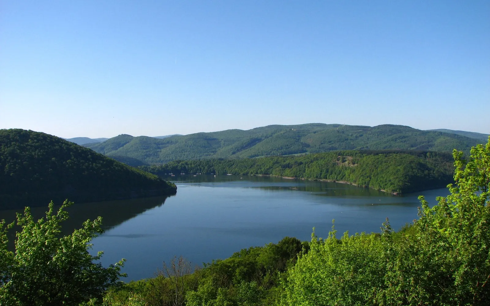 A wide view of a calm reservoir with tree-covered hills in the background under a clear blue sky.