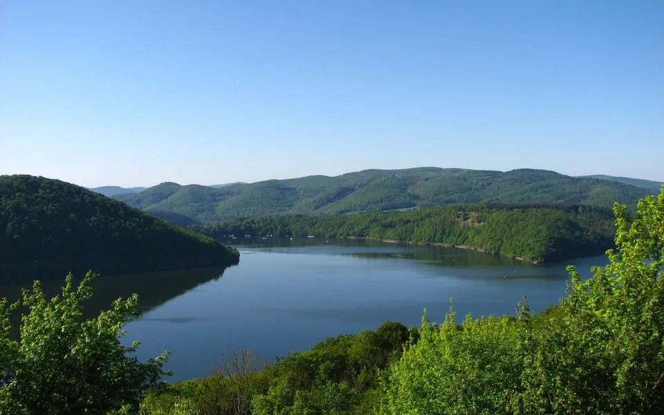 A wide view of a calm reservoir with tree-covered hills in the background under a clear blue sky.