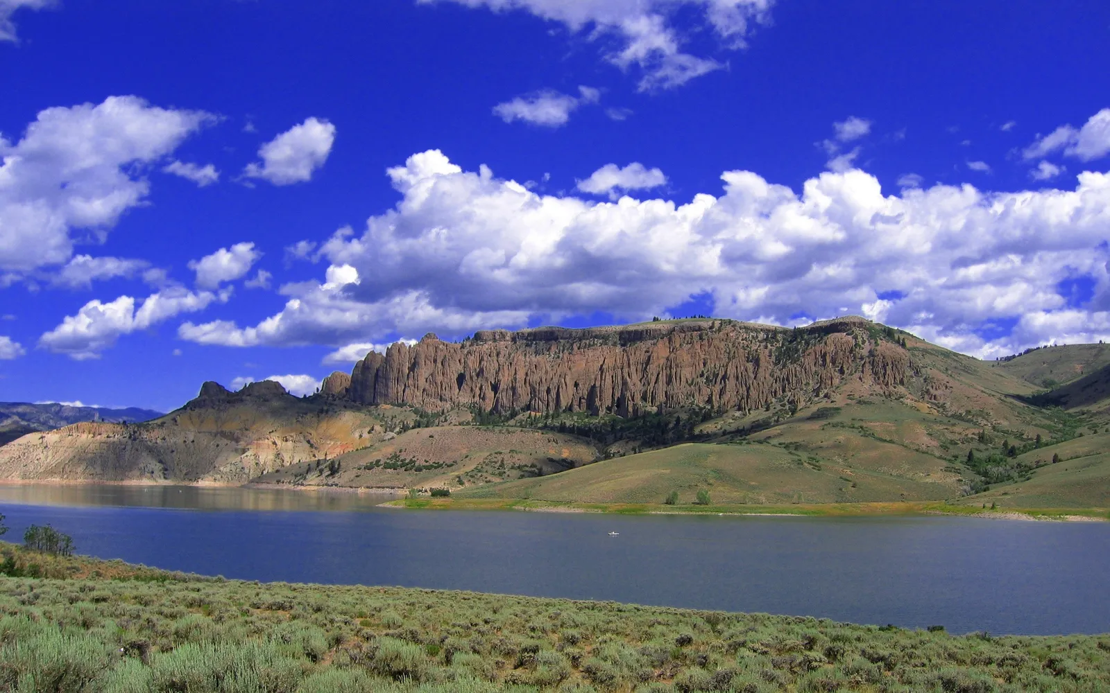 A wide, calm reservoir reflects the bright blue sky with scattered white clouds, with rugged, dry mountains in the background.
