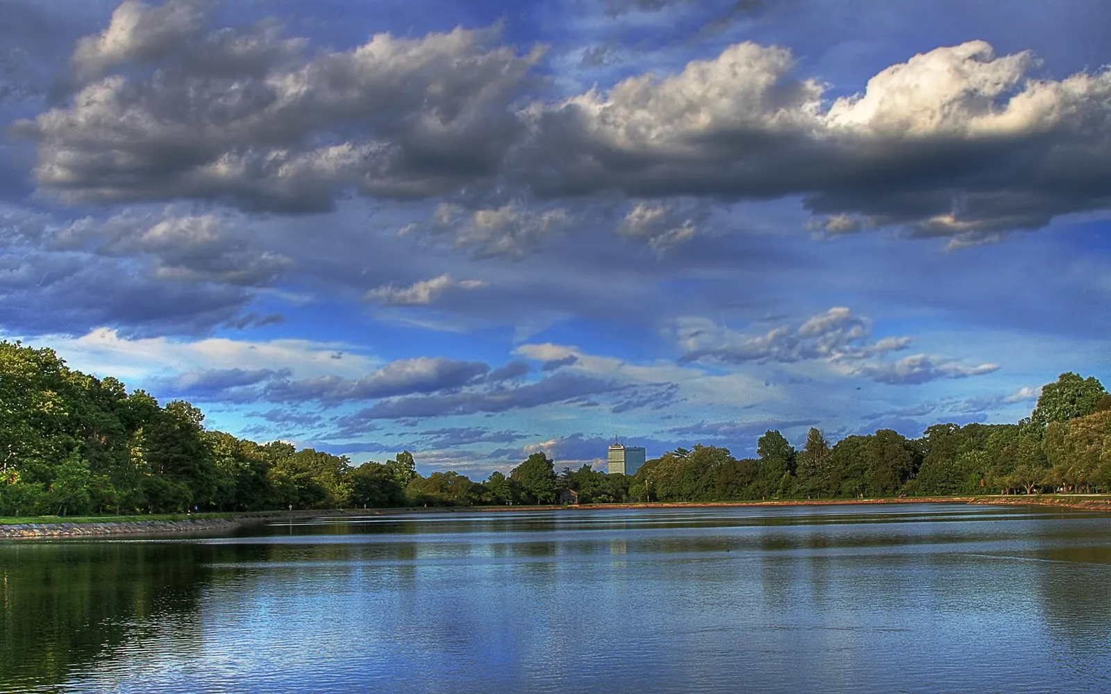 A wide, calm reservoir reflects the sky and surrounding trees under a dramatic, cloudy sky.