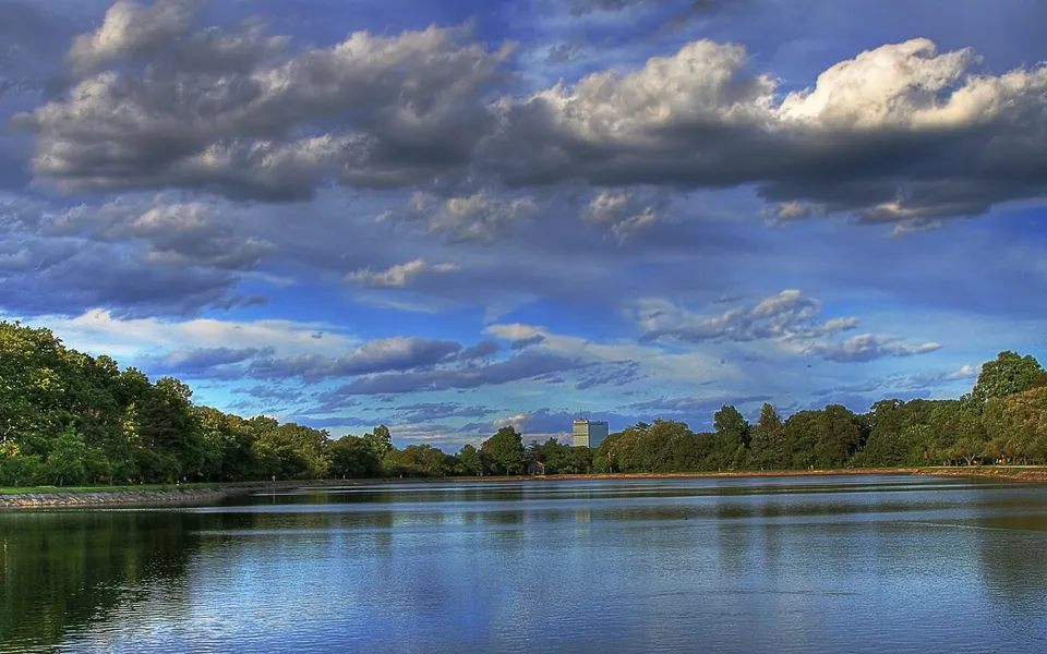 A wide, calm reservoir reflects the sky and surrounding trees under a dramatic, cloudy sky.