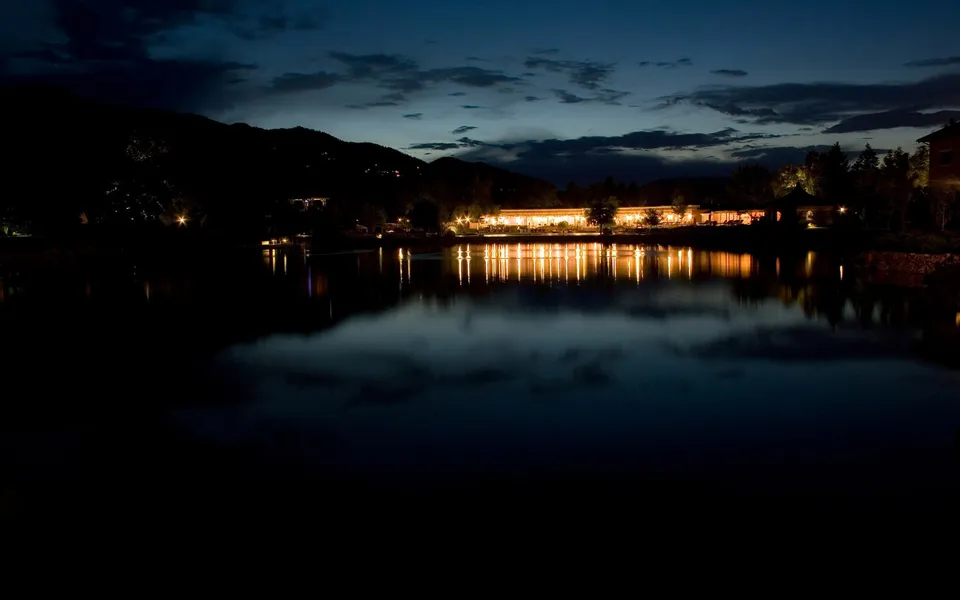 A long building with many illuminated windows reflects in a dark, still lake under a twilight sky.